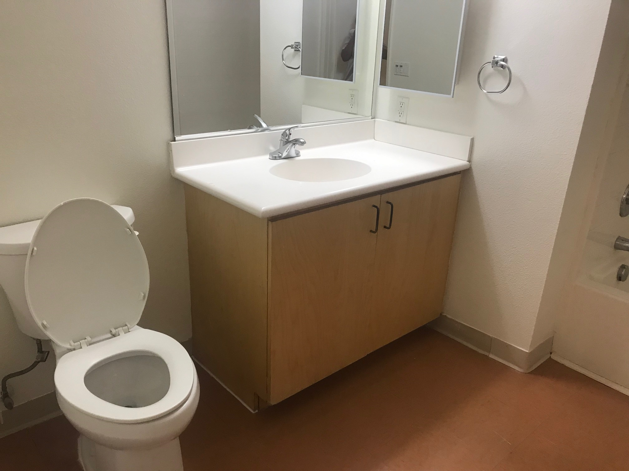 Angle view of a restroom with a white toilet, sink, two mirrors, small towel holder and lower cabinets under the sink.