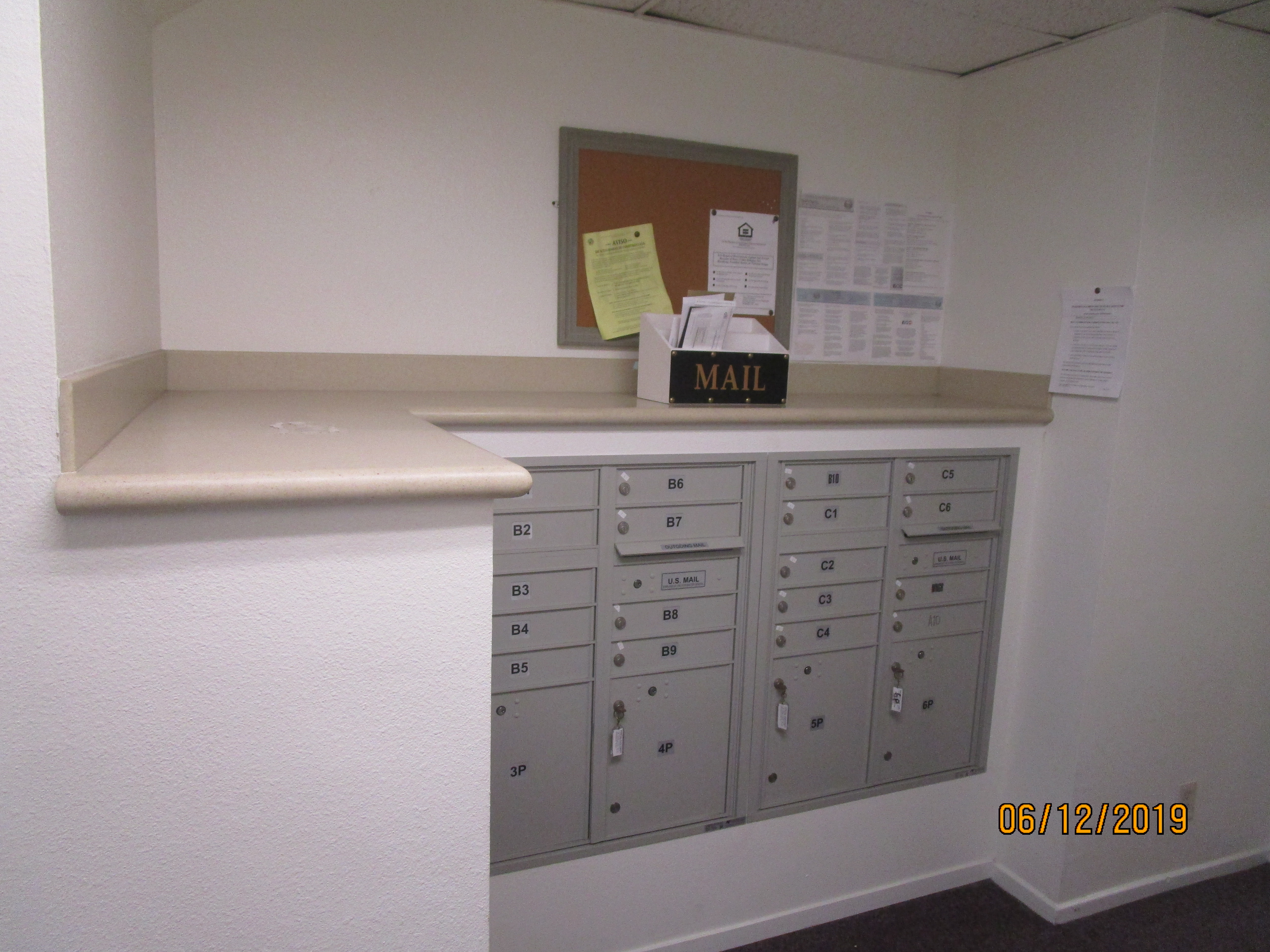 View of a mail room, a pin board on the wall, a white MAIL box, a set of gray locked mail boxes, Fair Housing flyers on the wall.