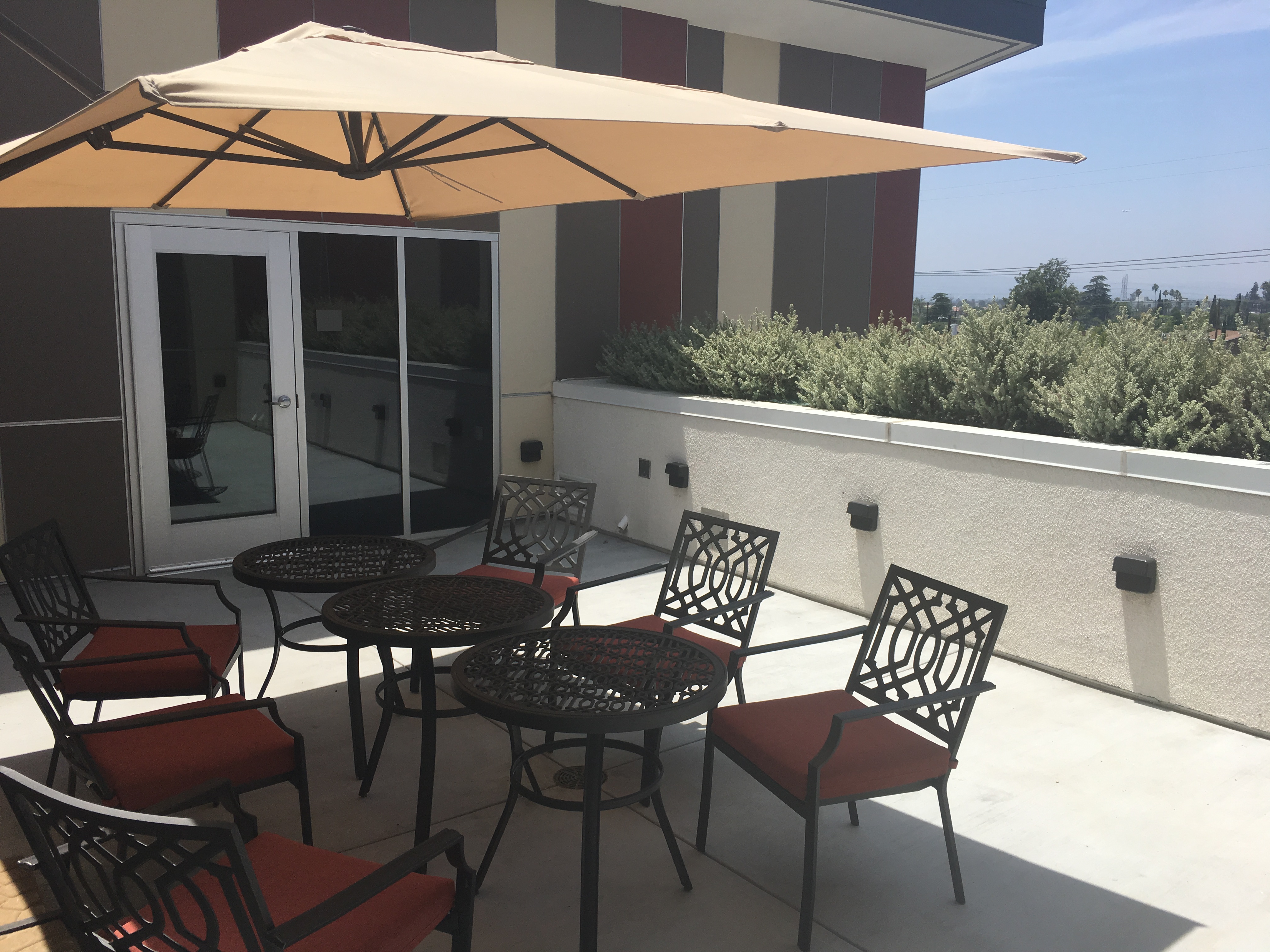 Exterior view of a balcony on the fourt floor in the PSH Campus. Three small round tables with six chairs and large open patio umpbrella.