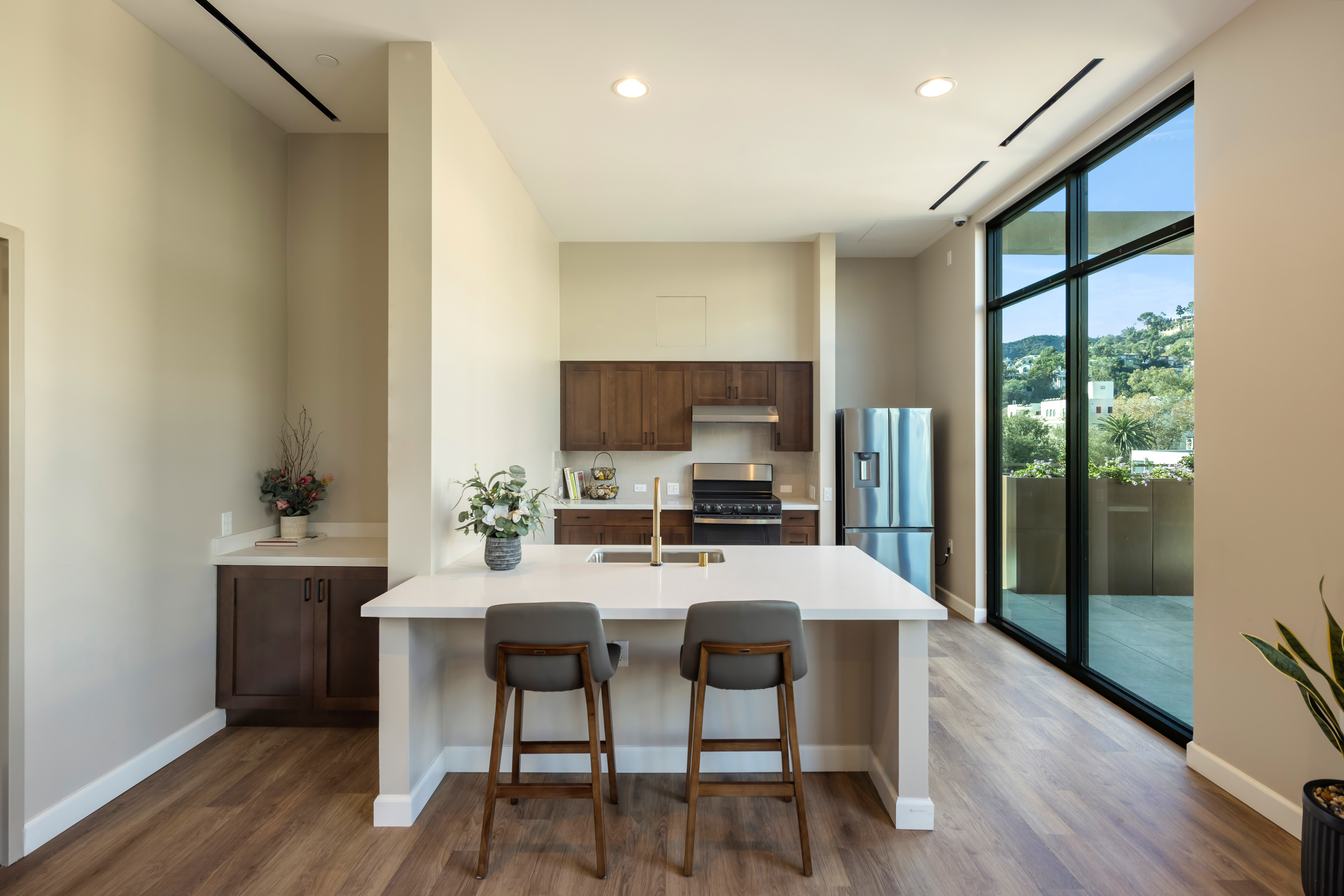 Modern community room with high ceilings and natural light from large windows. Features a central island with two stools, wooden cabinetry, and stainless steel appliances. Warm, inviting ambiance with plants and light wood flooring.