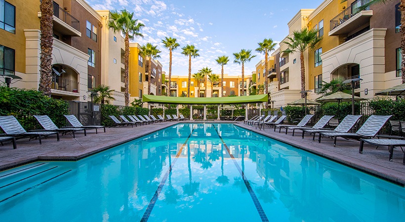 View of a very long pool, multiple chaise lounges around the pool, multiple units with balconies, tall palm trees.