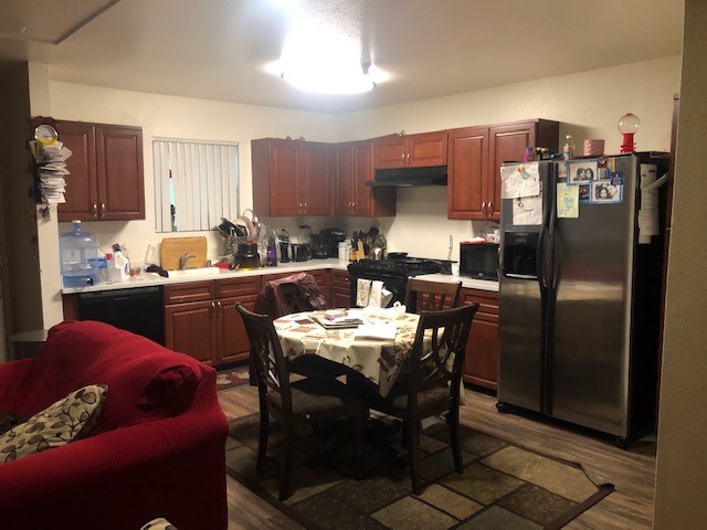 Inside view of a unit's kitchen area. Area contains a small dining set, a refrigerator, stove, dishwasher and microwave. Cabinets are brown and there are various items surrounding the sink like cookware. Unit seems to be occupied by a tenant.