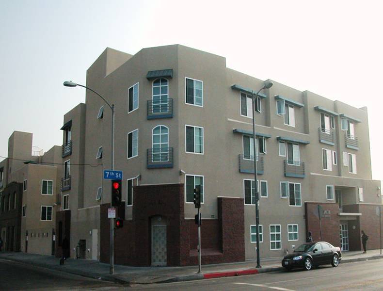 Photo of street view of a light brown four story building located on a corner intersection. 7th Street sign on lamp post.