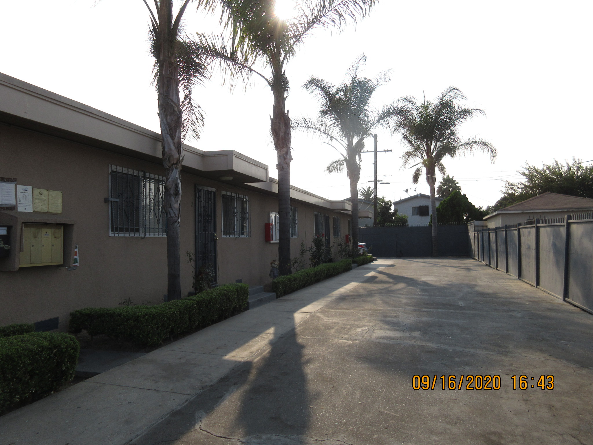 Outside view of a complex. There are mailboxes on the side with posted signs on top. Entry way to a couple of units are visible. There are small hedges in front of the units and trees. Windows have iron bars and doors have iron screens.