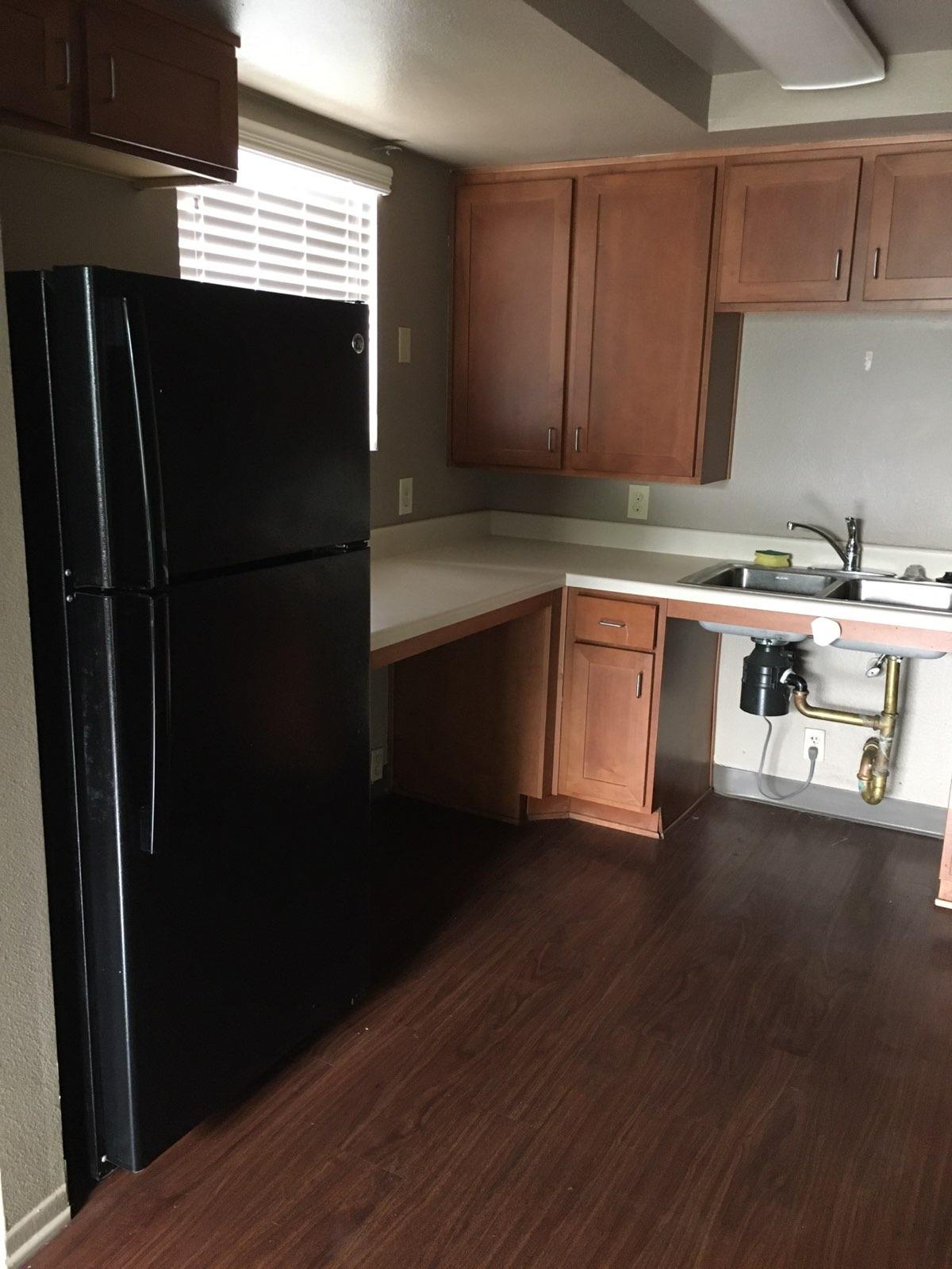 View of upper and lower kitchen cabinets, white countertops, black refrigerator, and wheelchair accessible sink.