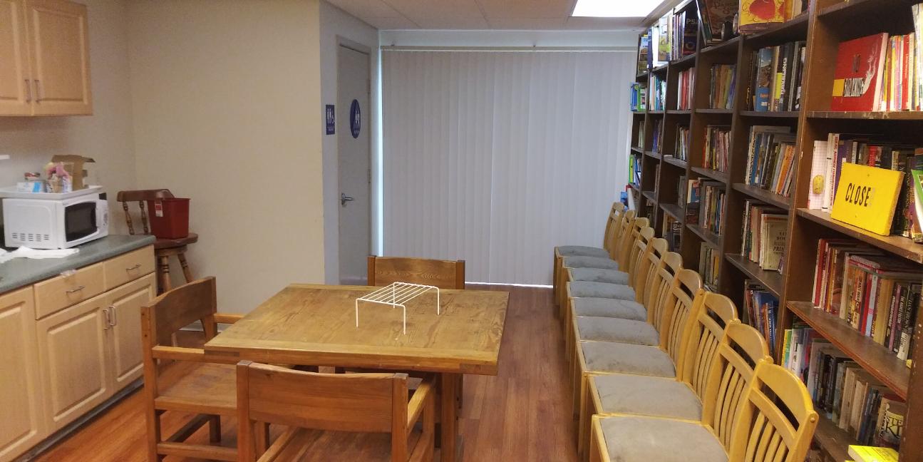Community room with a small dining table set, large bookcase with many books, and multiple chairs lined up against the bookcase. There is a microwave on a counter that has cabinets,, and a gender neutral restroom to the side.