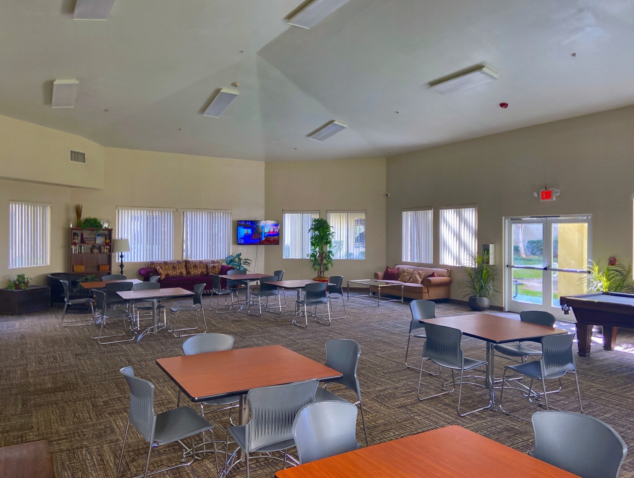 Large community room with square tables and chairs. There is a couch on one side of the room and a television on the other side of the room.