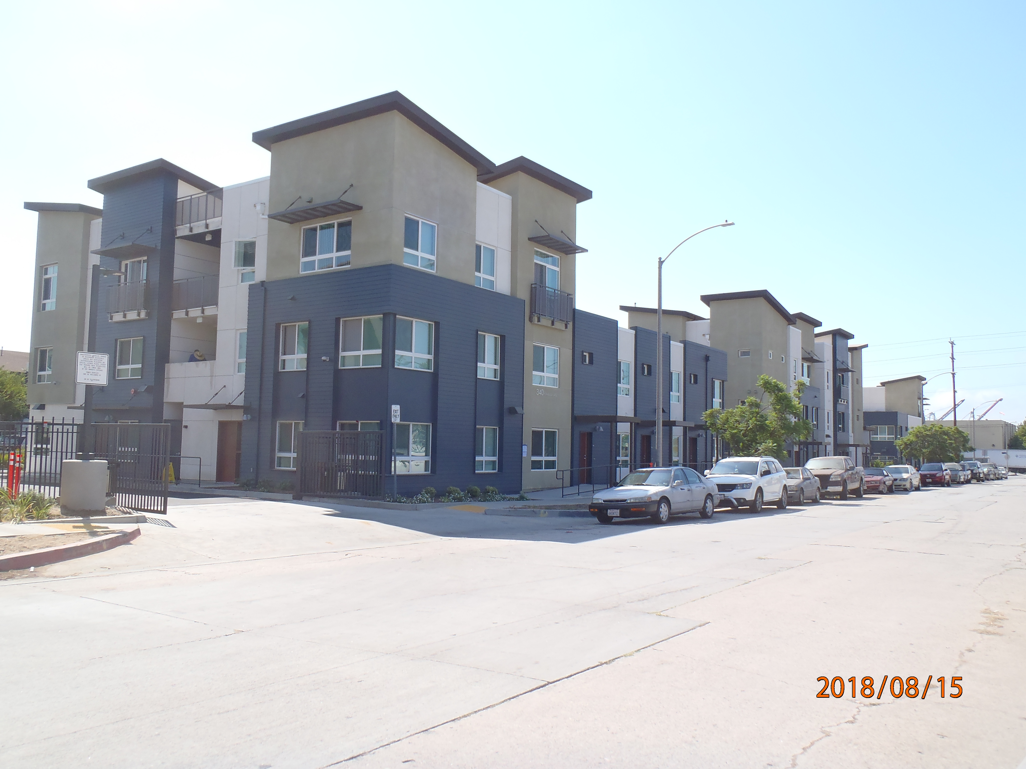 Corner view of a colorful block long three story building, multiple windows, concrete ramp with iron handrails toward the entrance, parked cars, open gates on the left side of the building.