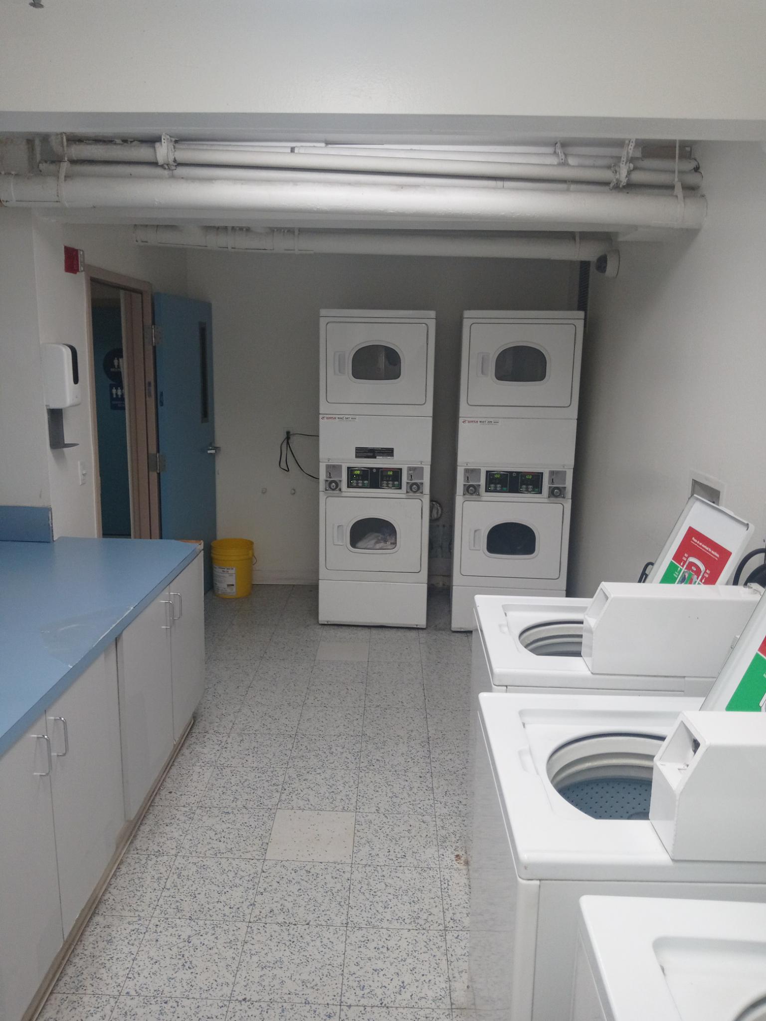Laundry room. Three washing machines, four dryers and a flat surface for folding are visible. Blue door is propped open.