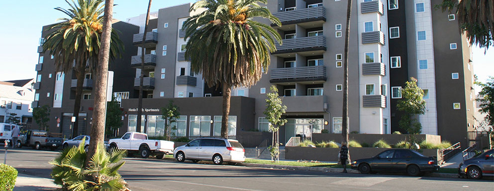 Street view of Menlo Family Housing. Modern styled Multi-level building painted with neutral tone colors. Building has balcony access from the units. Stairs and accessibility ramp leads up to building entrance. Large palm trees along building front and la
