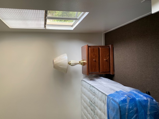 Right side view of inside a white room, new set of mattresses, brown two drawer nightstand, beige lamp, window with a white horizontal blinds, brown carpet.