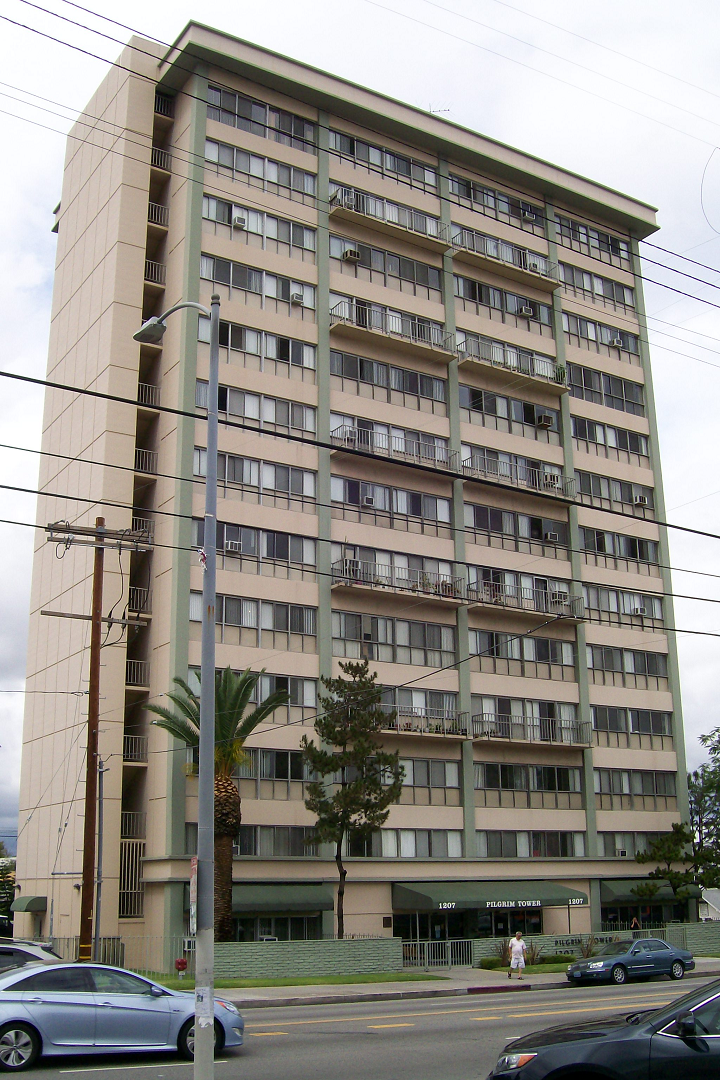 Street view of 13 floor tan with green trim building with balconies, ac units and gated entrance