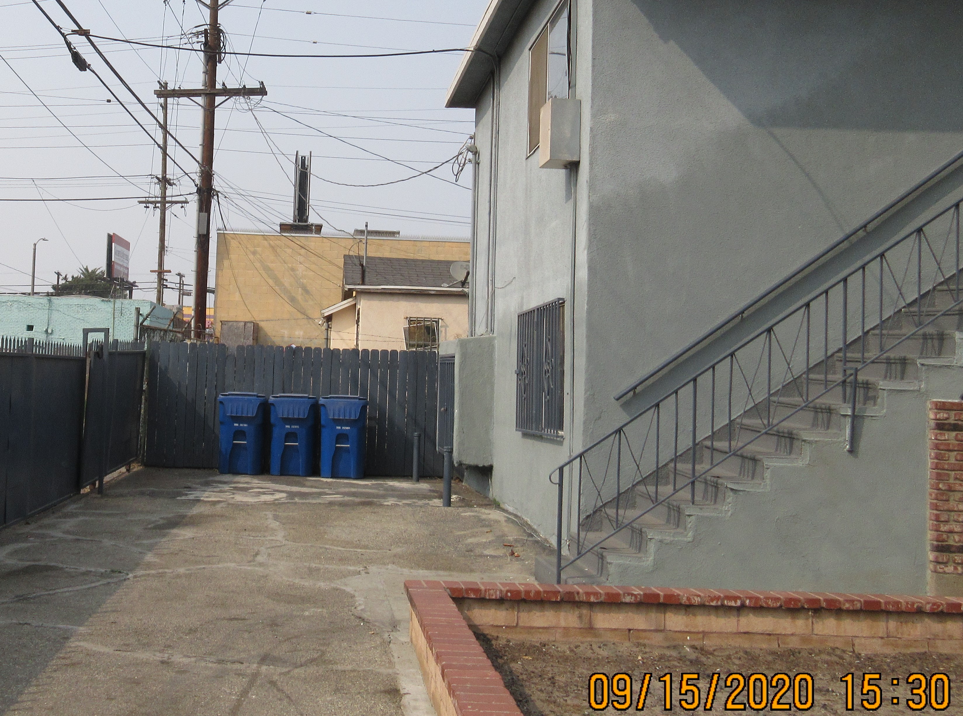 There is a 2 story blue building on the right with a set of stairs leading to the second floor. View of patio, three recycle bins.