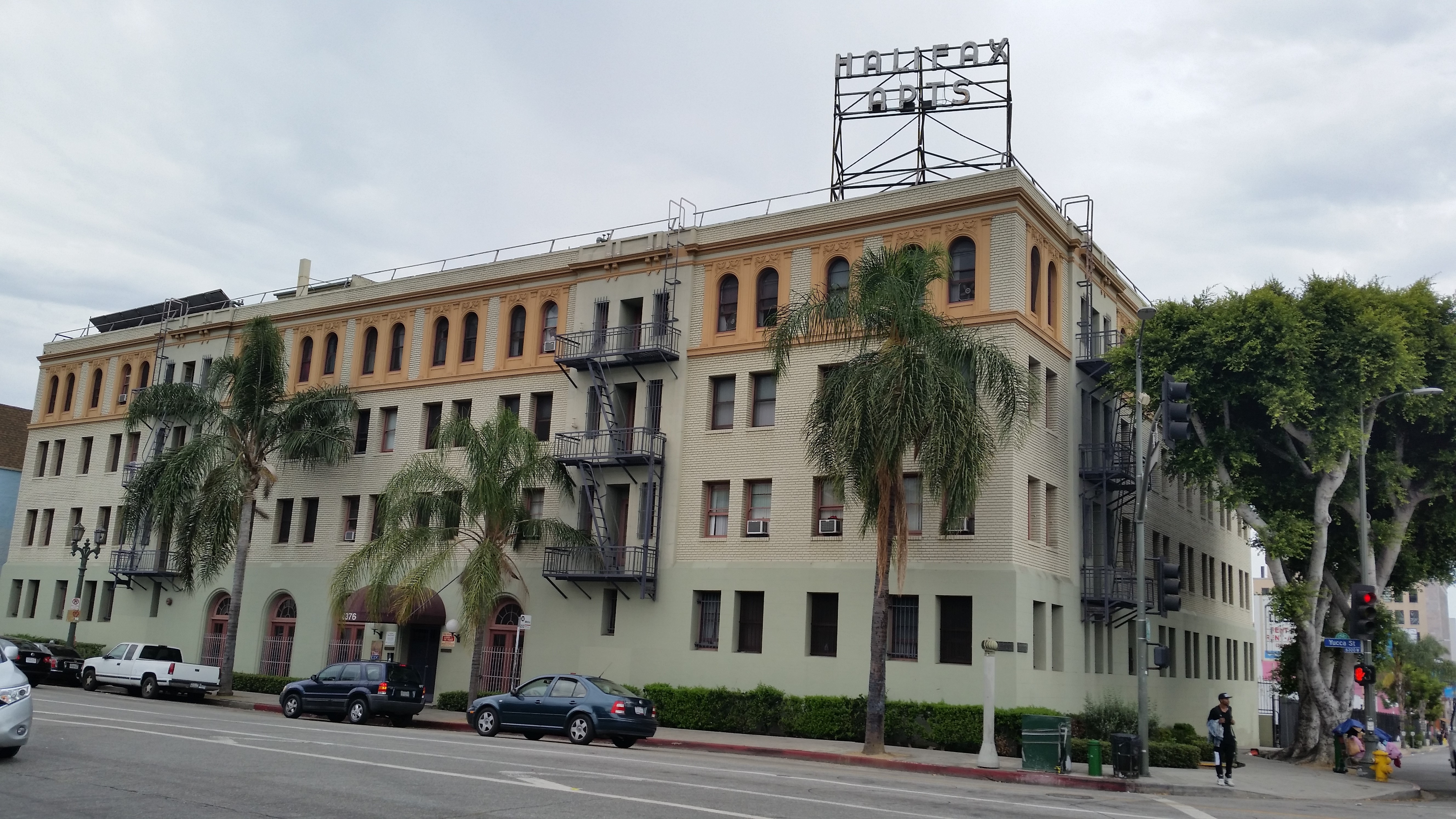 Street view of 4 story white, brick building with fire escapes and hedges along building, some units with a/c, some windows have bars, strre parkingin front of building