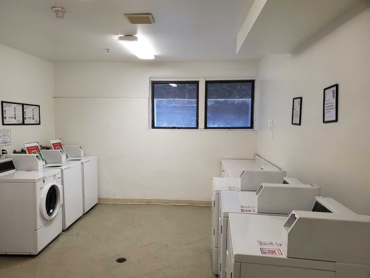 Laundry room with multiple washer and dryer machines across from each other. There are two small windows and some signs posted on the wall.
