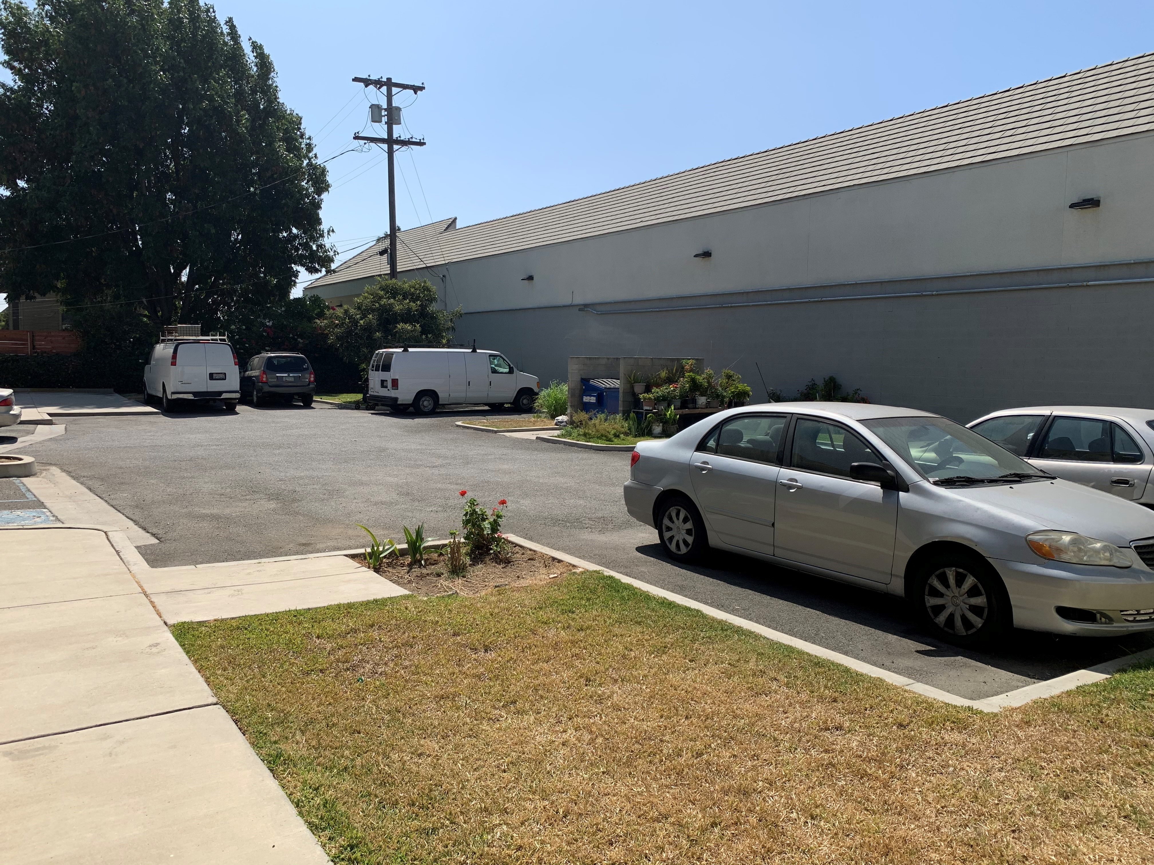 View of an outdoor parking lot. There is a dumpster area in the middle back. And there are sections of grass area and plants surrounding the lot.