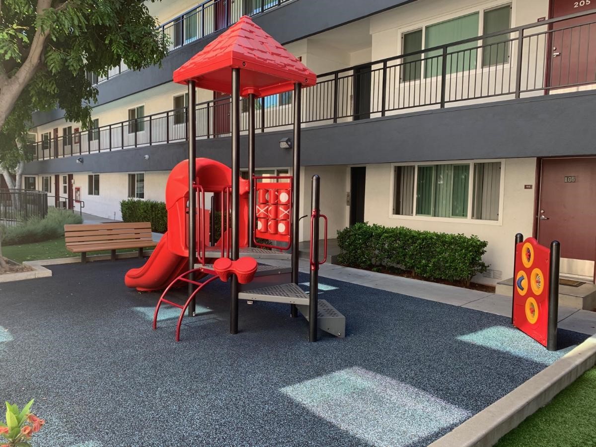 Right side view of a red playground, gray soft pave flooring mat, wood bench on left side, bushes around the playground.
