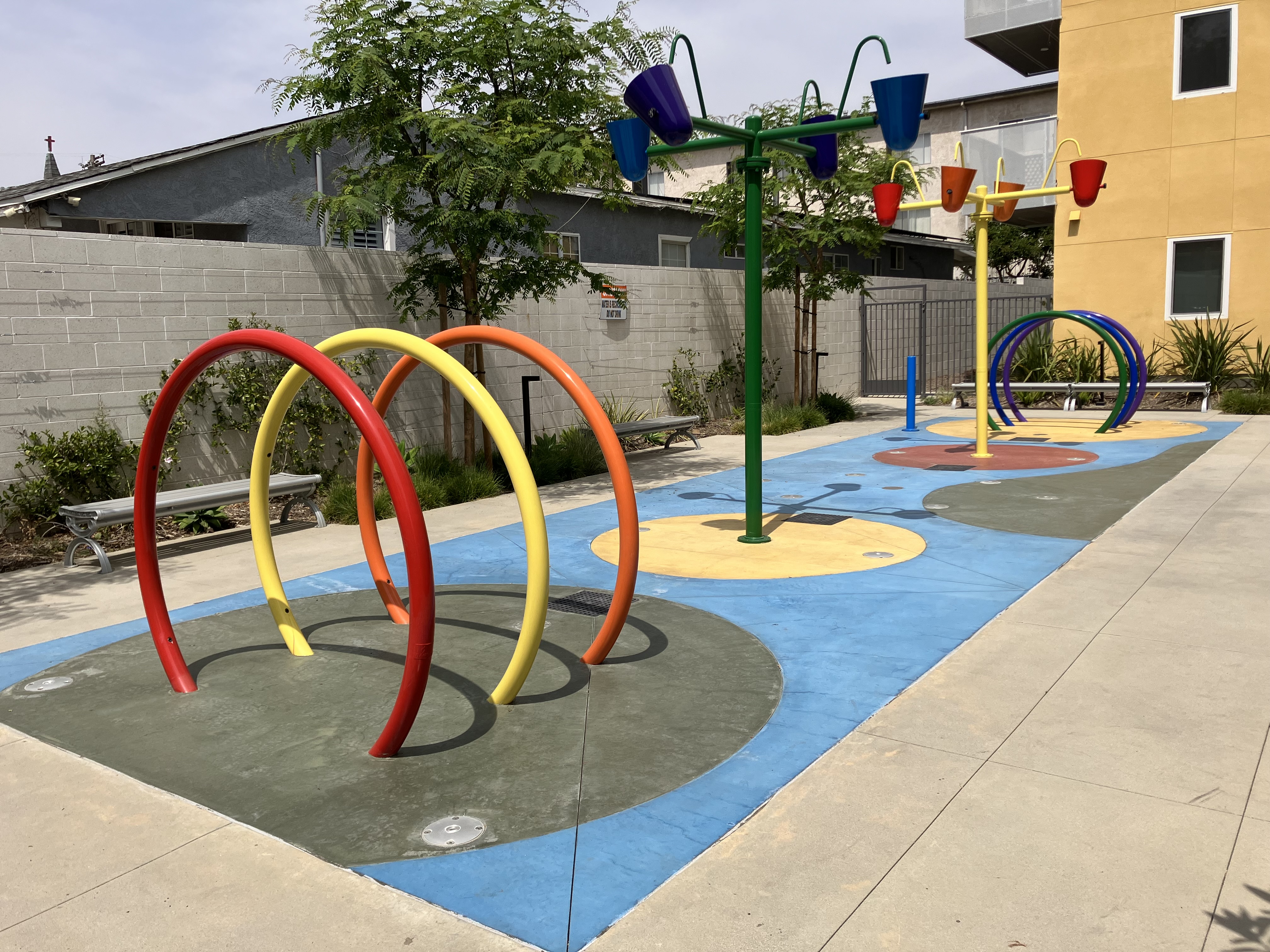 Mission Gateway Splash Pad Area. There are benches and trees surrounding the area. The Splash Pad Area includes playground equipment.