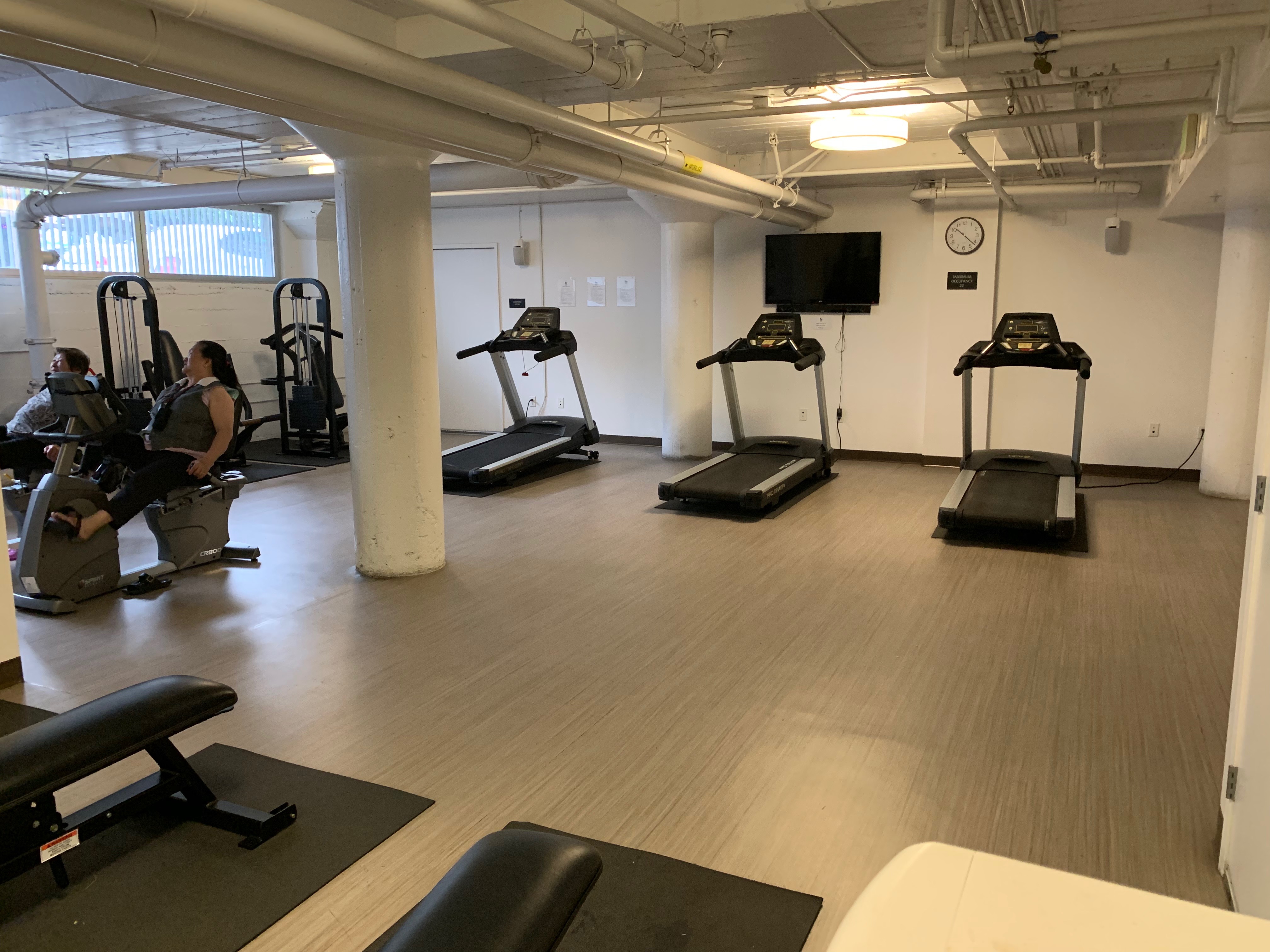 Fitness room that consists of a couple of treadmills, bench presses, weight stack machine and excercise bike machine. There is also a flat screen and clock on the wall.