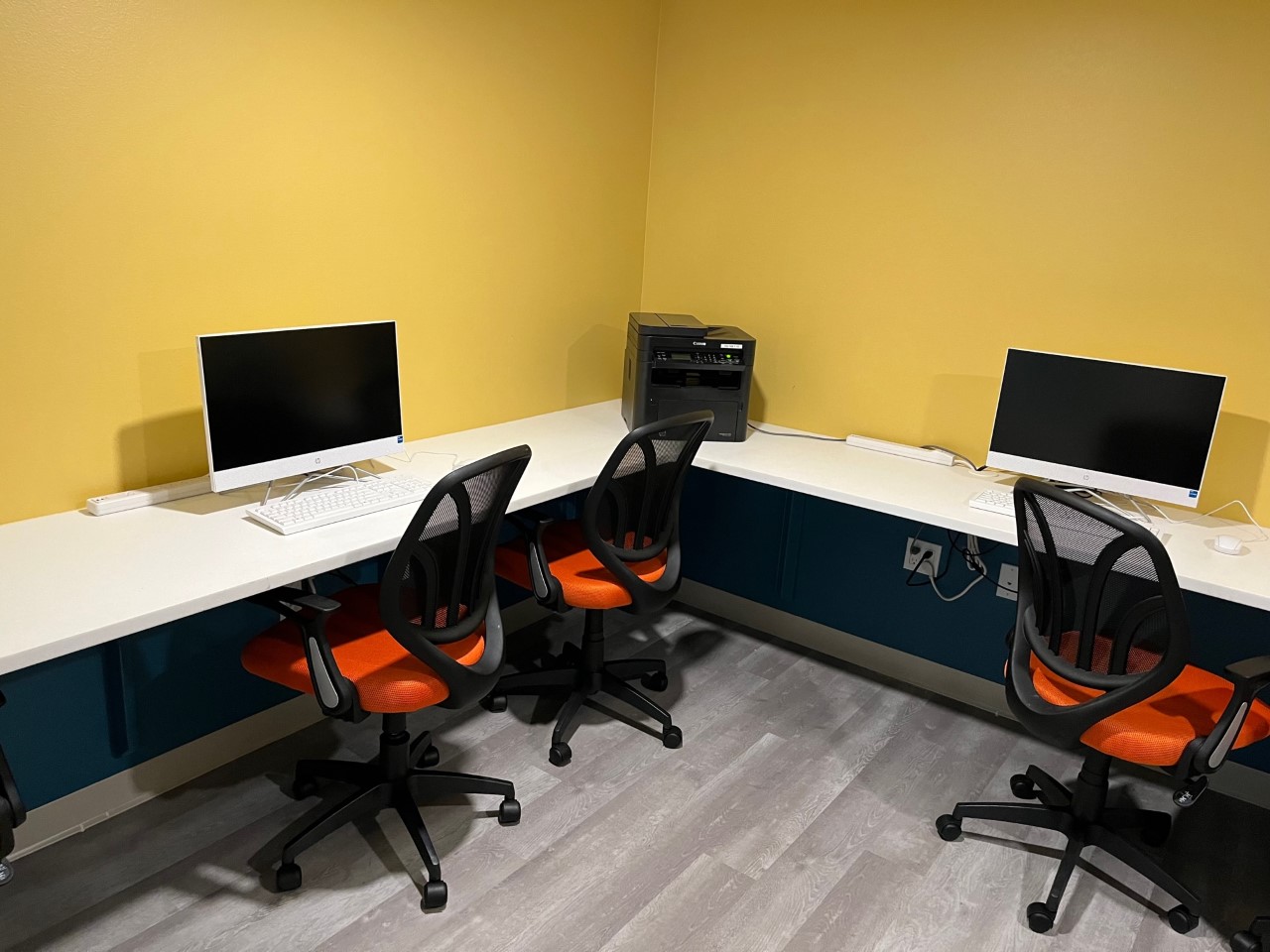 A room with blue and yellow walls. Two computers on a white desk with three office chairs.