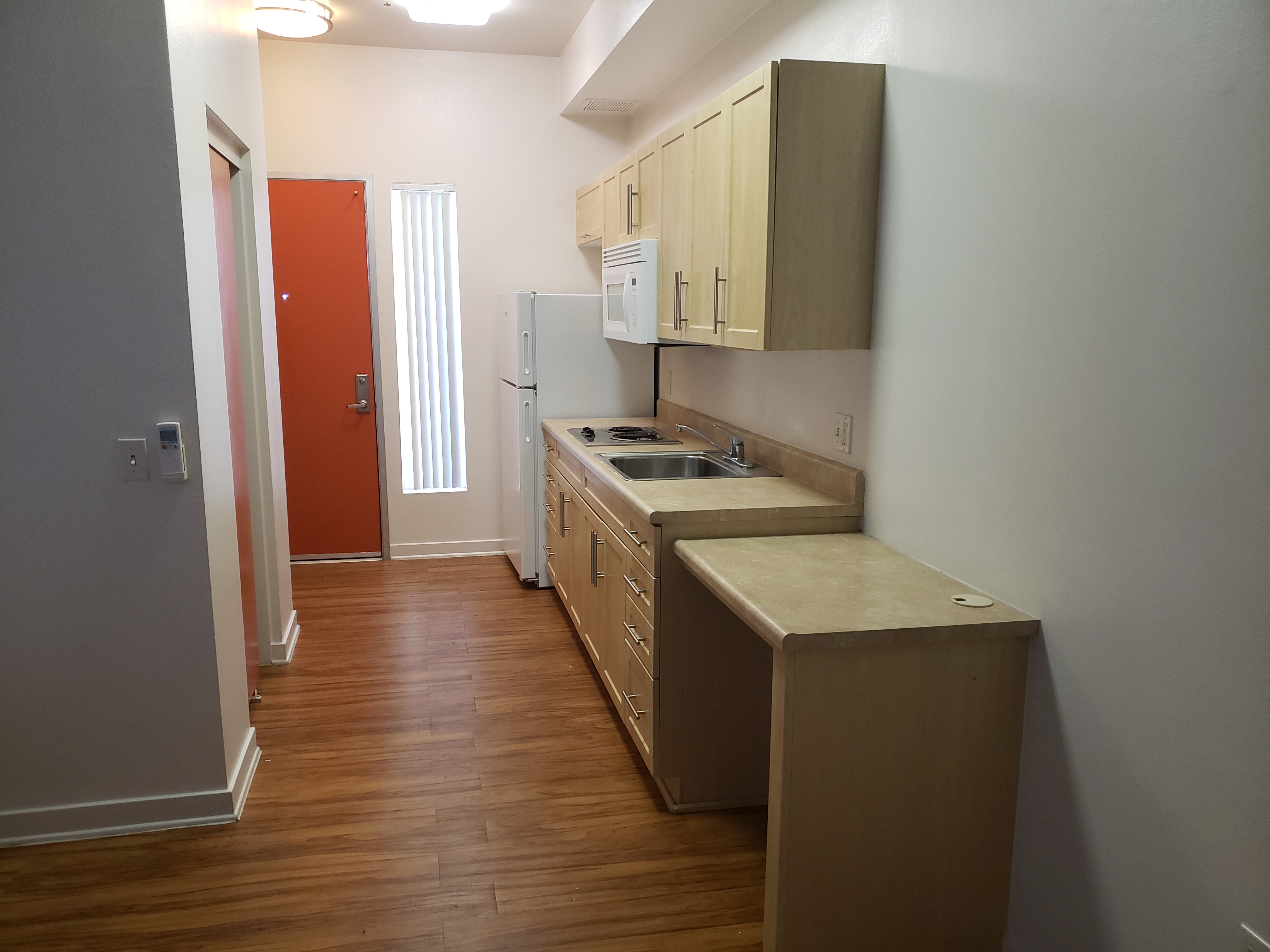 view of walk in kitchen. Upper and lower cabinets, fridge, two burner electric stove, large sink, and microwave attached to bottom of upper cabinets. At the end of the cabinets there is an attached wheelchair accessible table