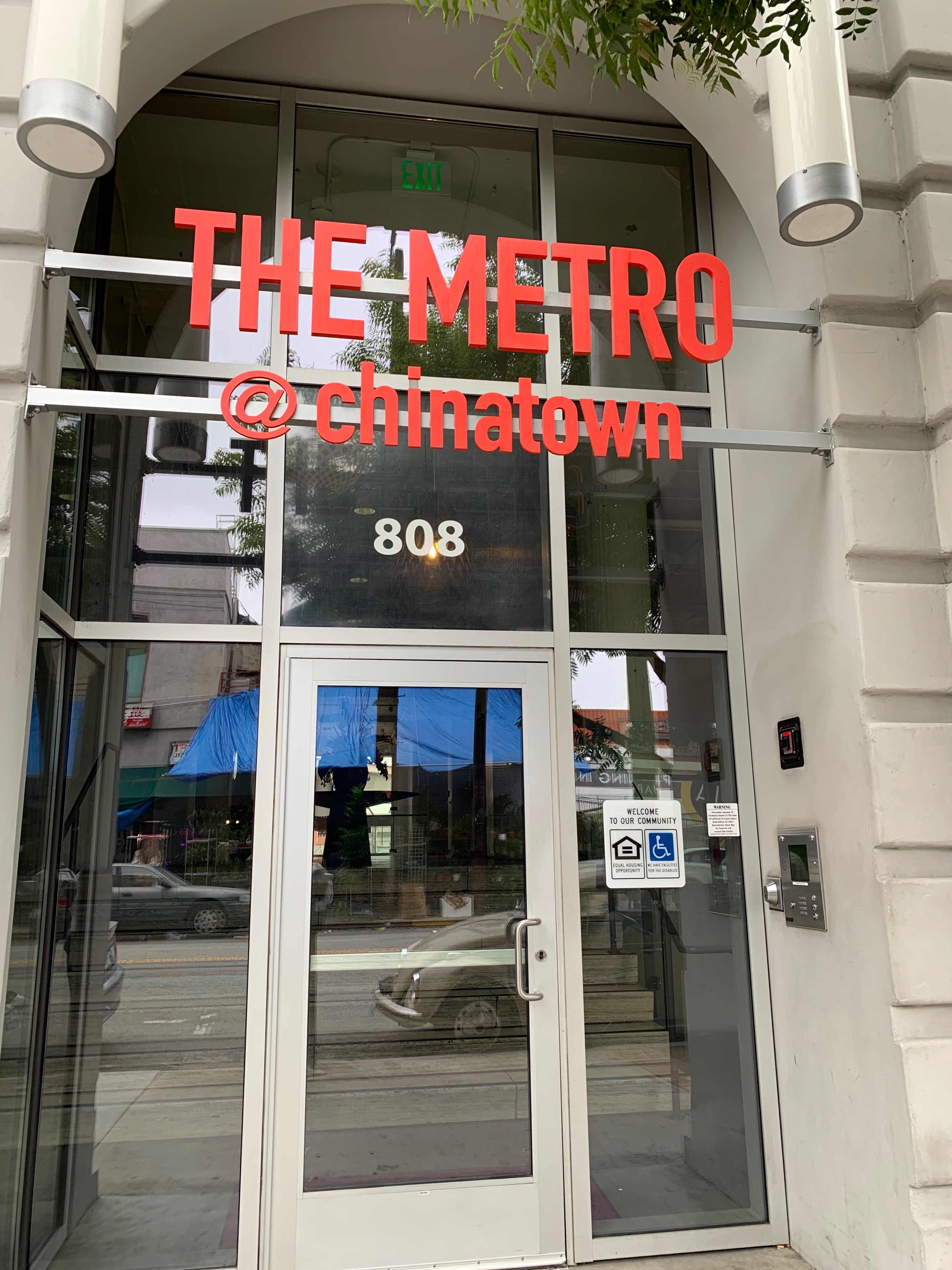 Front view of building entrance. Entrance consists of large windows and a glass door. There is a key pad on the side, and red letters read "The Metro @ chinatown" as a header above the door.