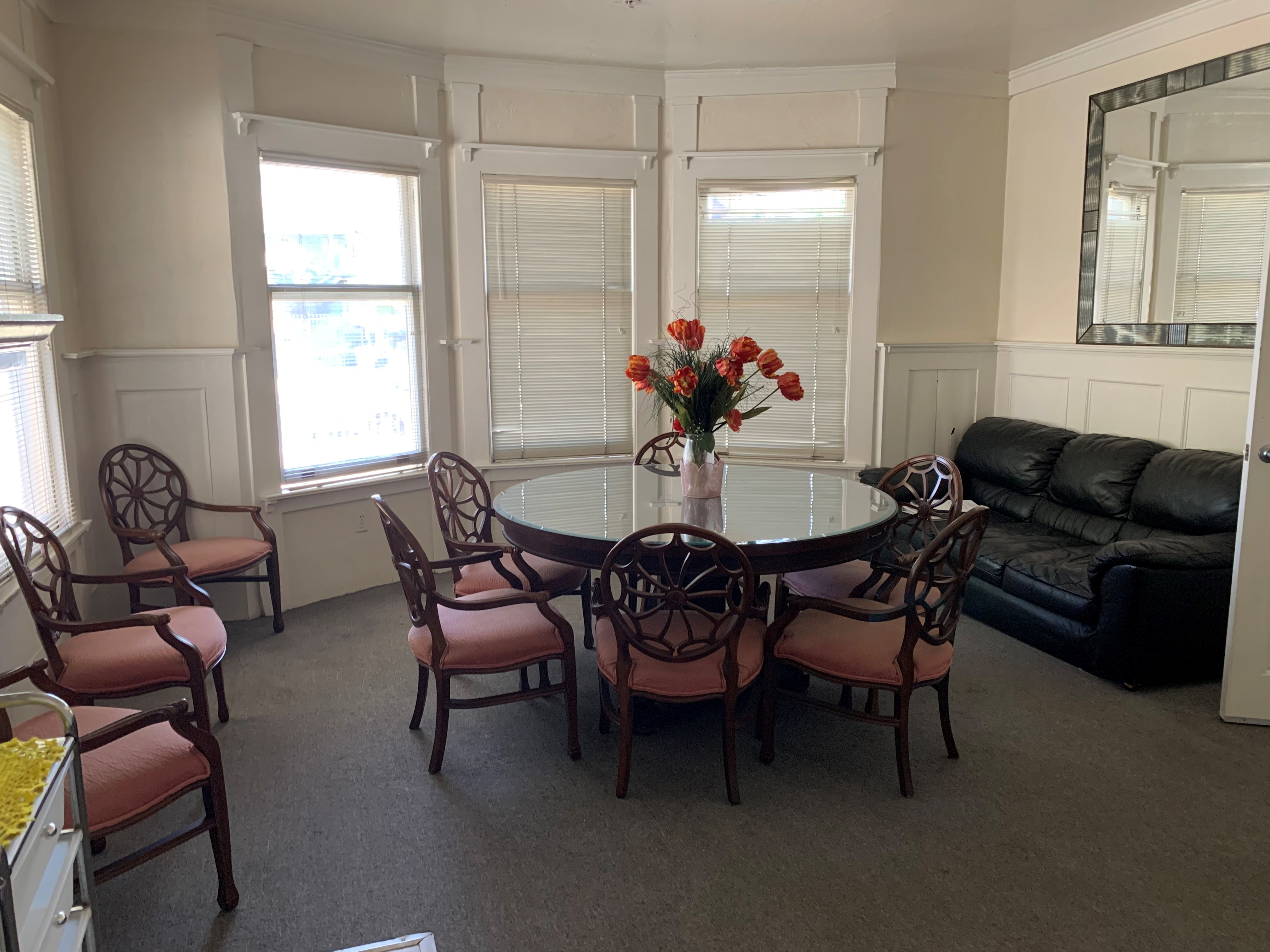 Lounging area with a round table in the center, chairs, and a sofa. There are four windows with blinds, and a mirror on the wall.