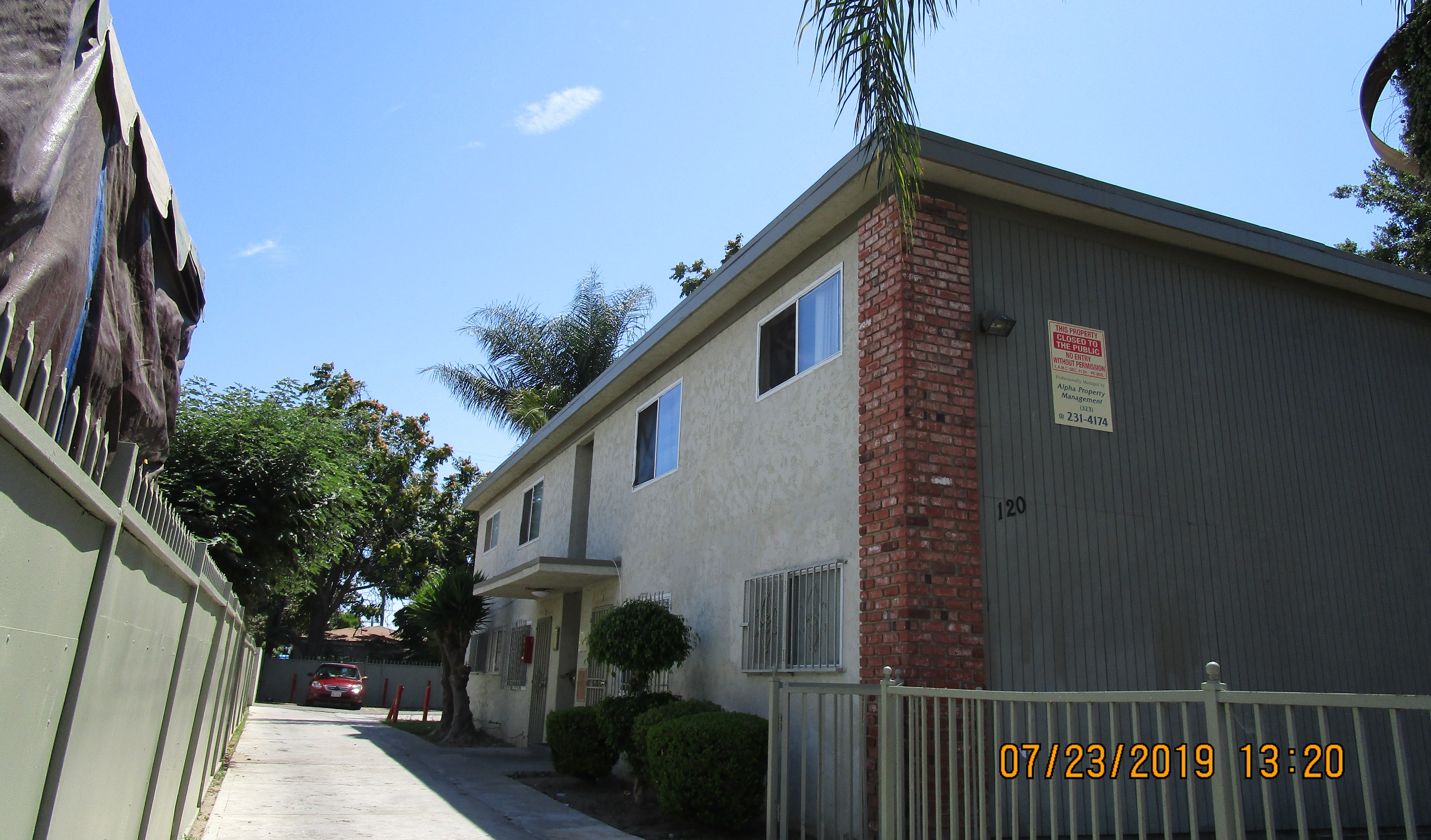 Left side of a beige two story building over looking the driveway, multiple windows in both floors, gated windows on the ground floor, iron screen doors, trees and bushes next to the units, a parked car inside the parking.  Open gate, closed to the public.