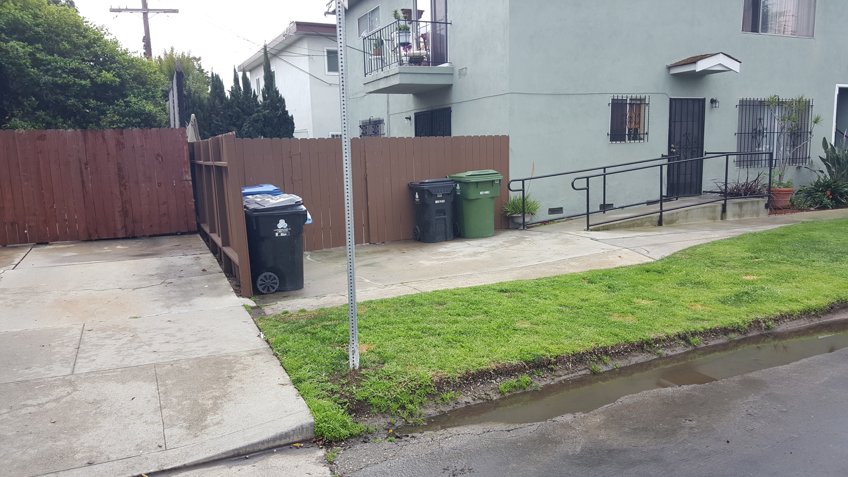 Image of 2 floor building with grass area in front yard. Lower level has a ramp leading to the front door, brown fenced in patio, windows with bars. Upper level has a balcony