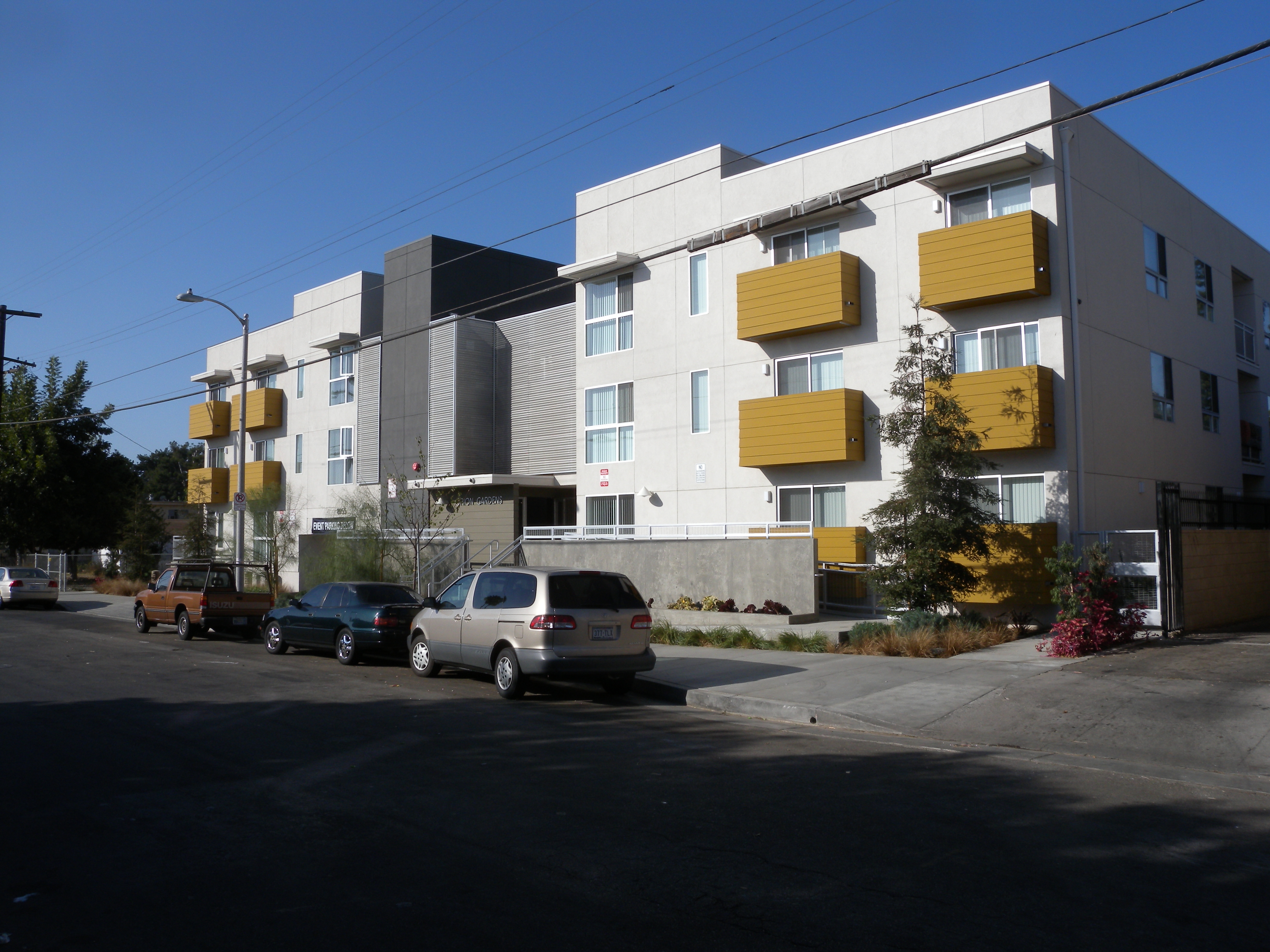Three story white and gray modern building. There is ramp and stairway access for entry way. Building has mustard yellow balconies for units that face the front.