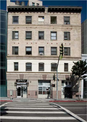 Five story light brown vintage building. Entrance is ground level and directly in front of a crosswalk. Top level has canopies.
