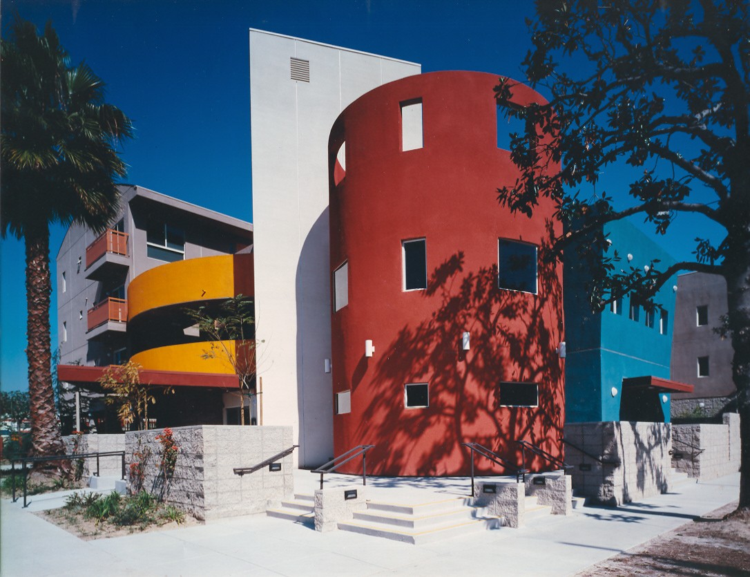 Corner view of a four story multicolor building. There is a wide four step staircase with handle bars. There are trees and plants on the sides of the building.