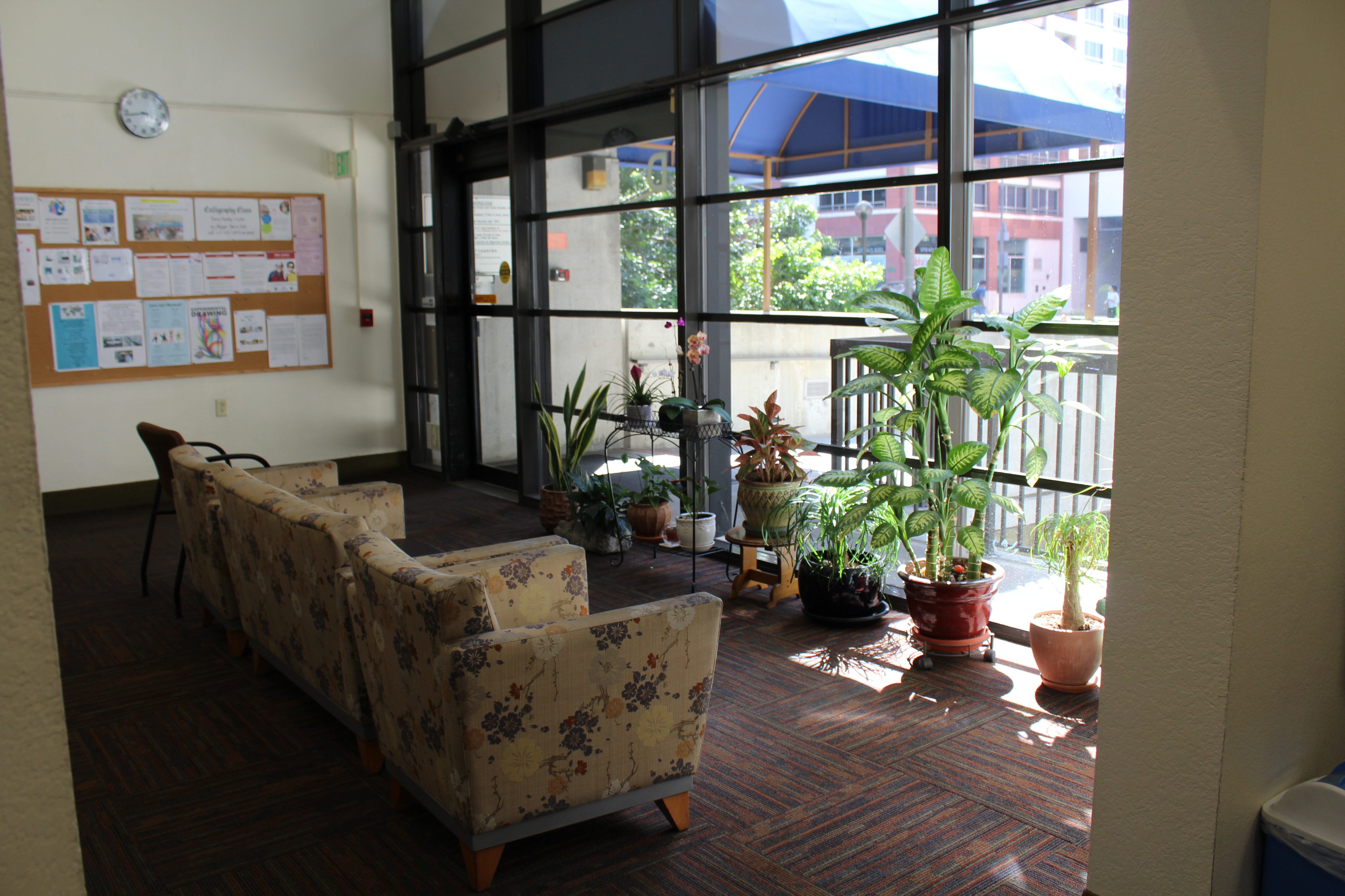 Interior view of a community area at Angelus Plaza 2. A few sofa chairs facing the windows, potted plants along the windows and a bulletin board with information attached.