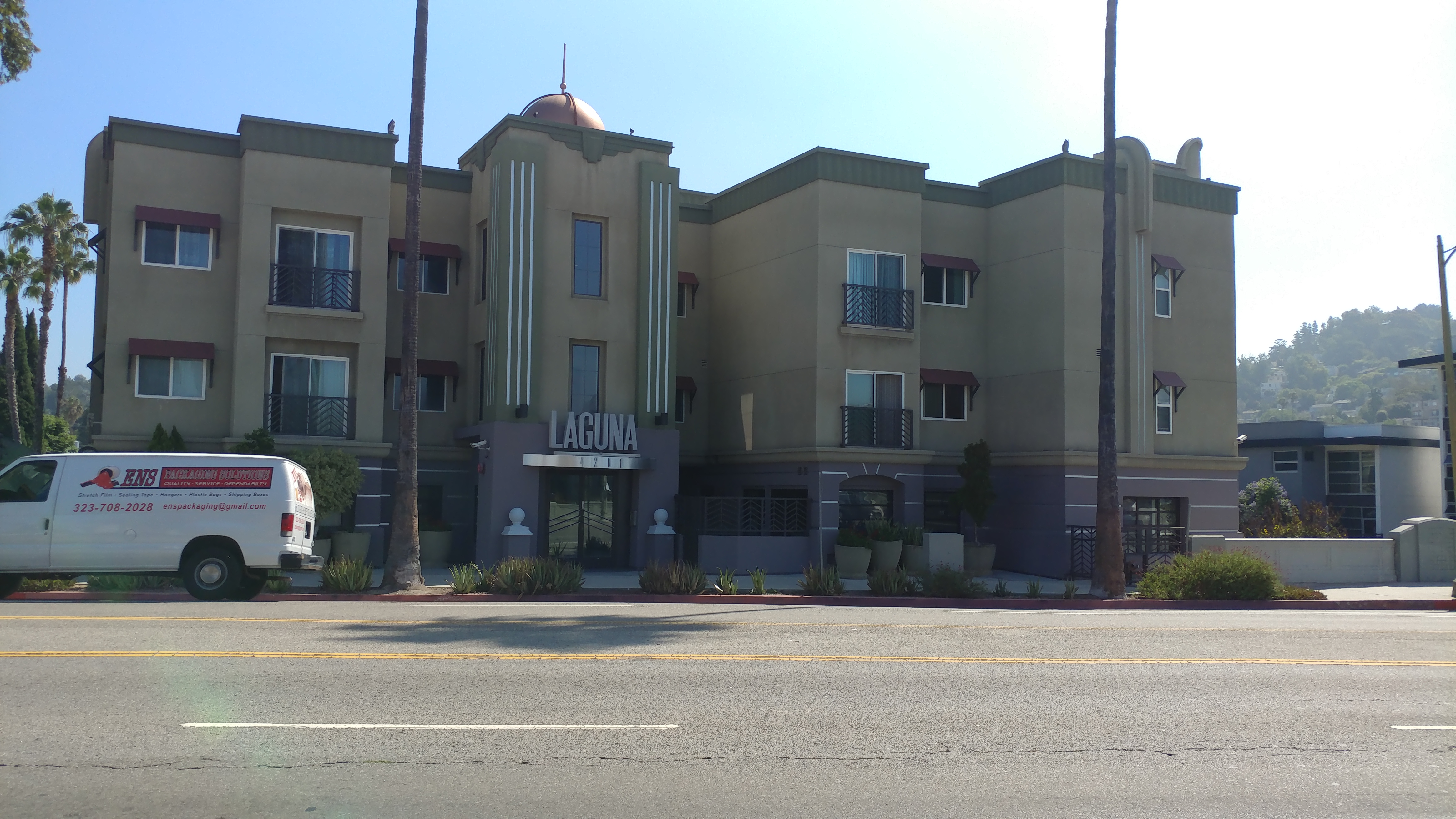 Front view of Laguna three story building, white van parked outside, multiple windows, sliding doors with guard rails on the second and third floor, big beige planters, tall palm trees.