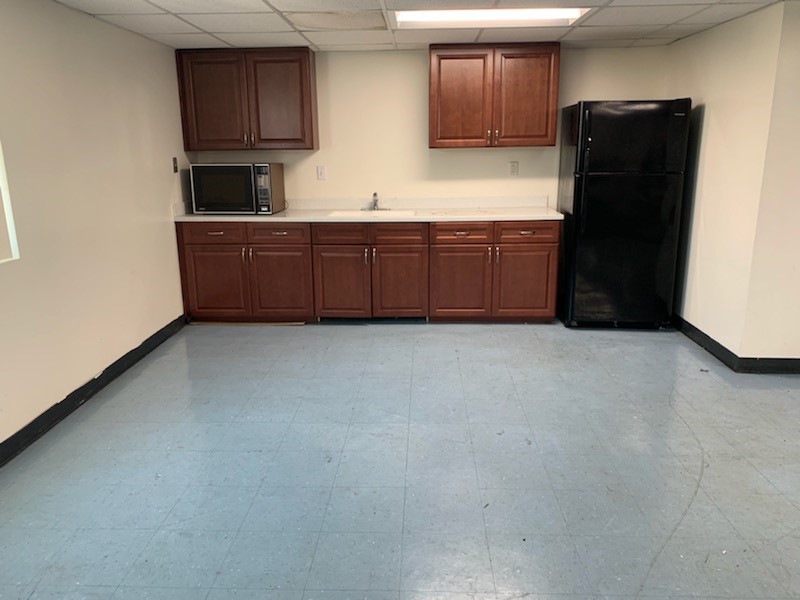 Image of a kitchen space. Area consists of a refrigerator, sink, microwave, and large brown cabinets. The front floor is spacious.