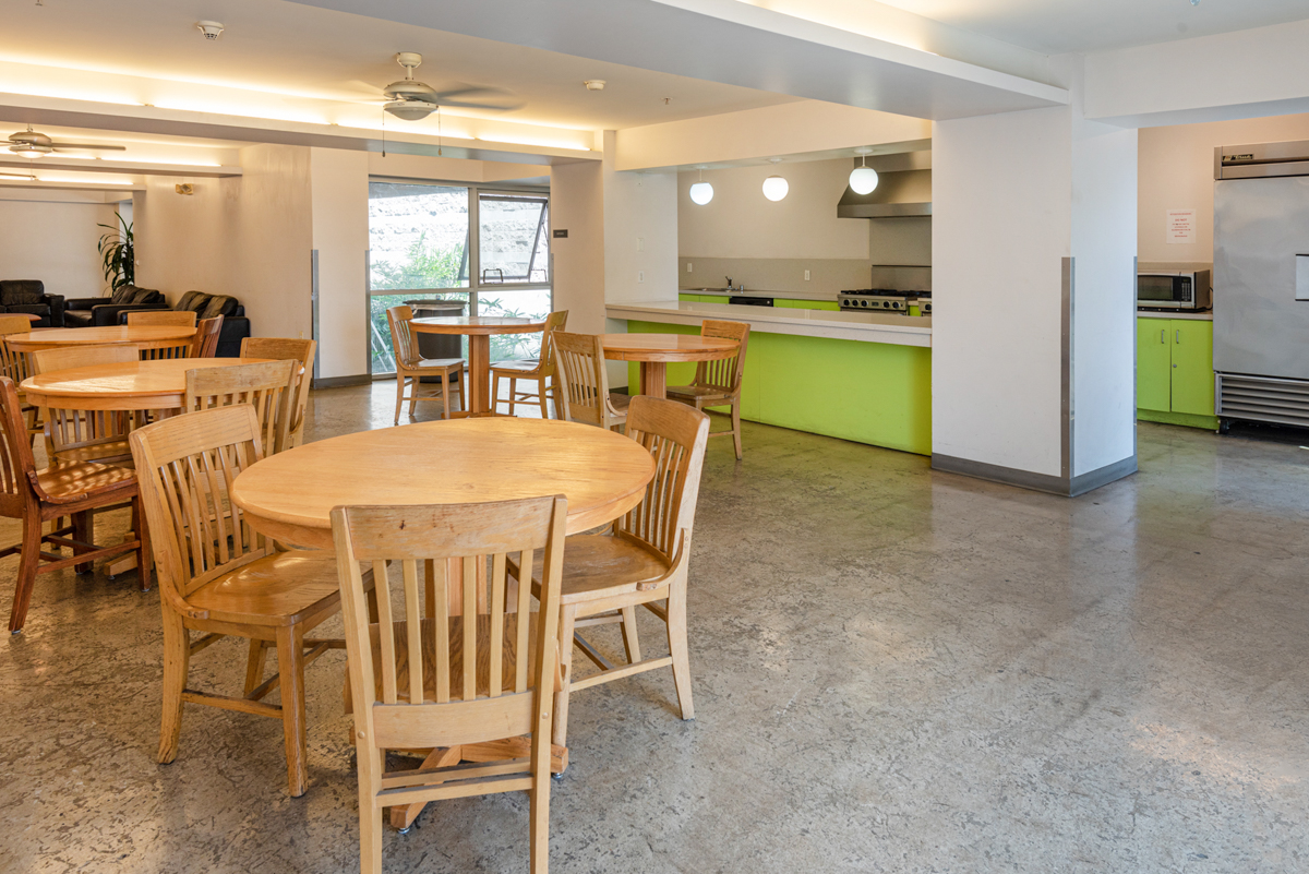 View of the kitchen, five round natural wood color table with chairs, a bar table, lime green pantry, sink, stove with oven and a top range, microwave, a commercial refrigerator, ceiling fans, three round decorative lights hanging from the ceiling.