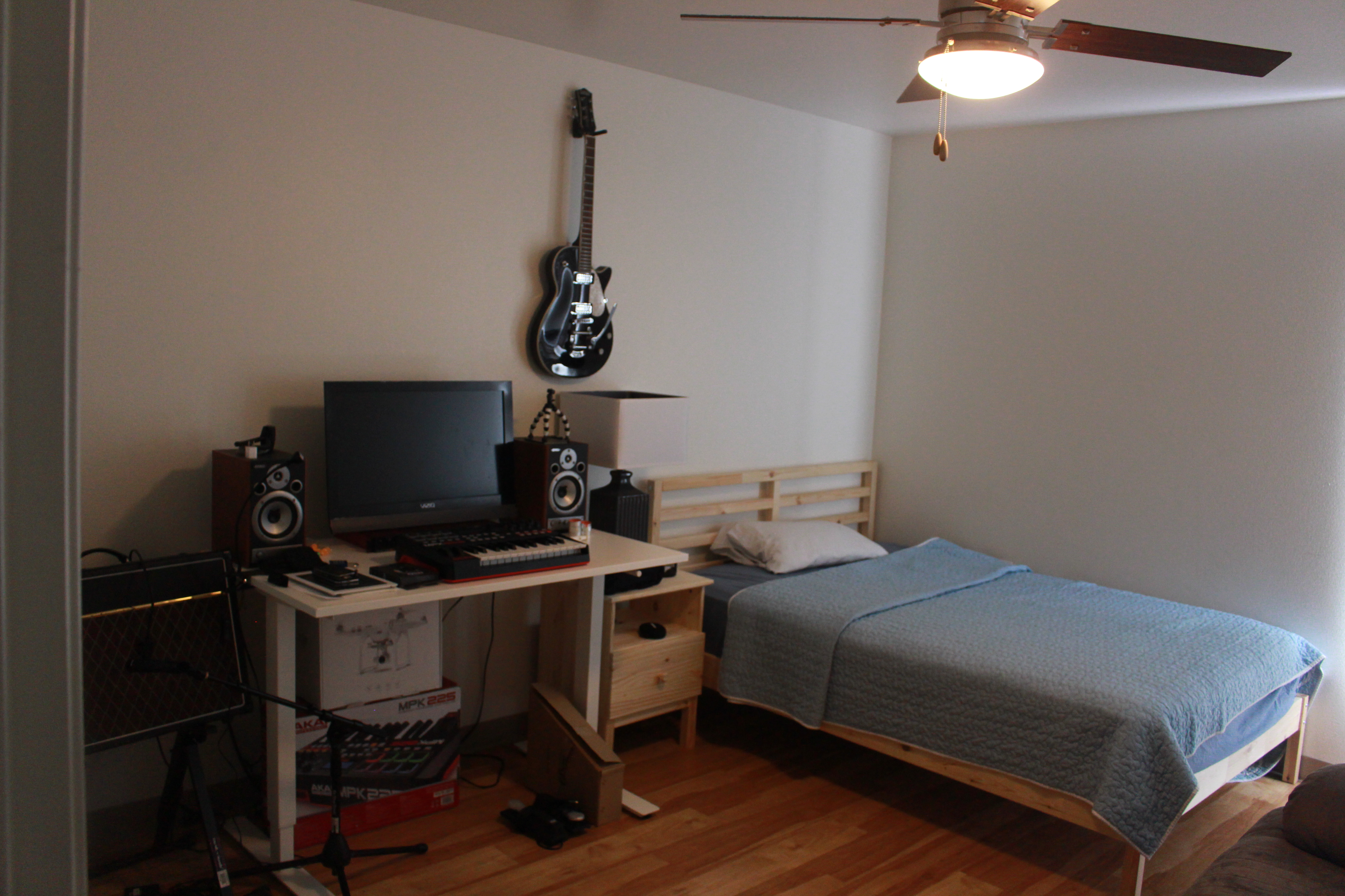 Beige wall room, laminate wood floors, a desk with a computer, a guitar and speakers and other music boxes, a modern black and white lamp on a natural wood bedside table, natural wood twin bed with a light blue comforter, white pillow, a ceiling fan with