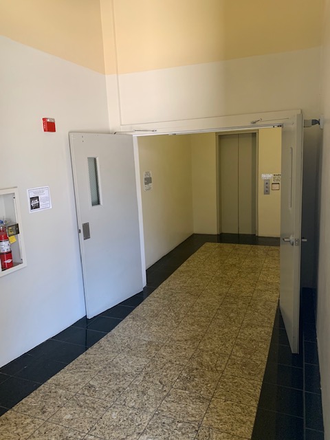 View of a lobby, double gray wide open doors, elevator, fire extinguisher, two color tile floor.