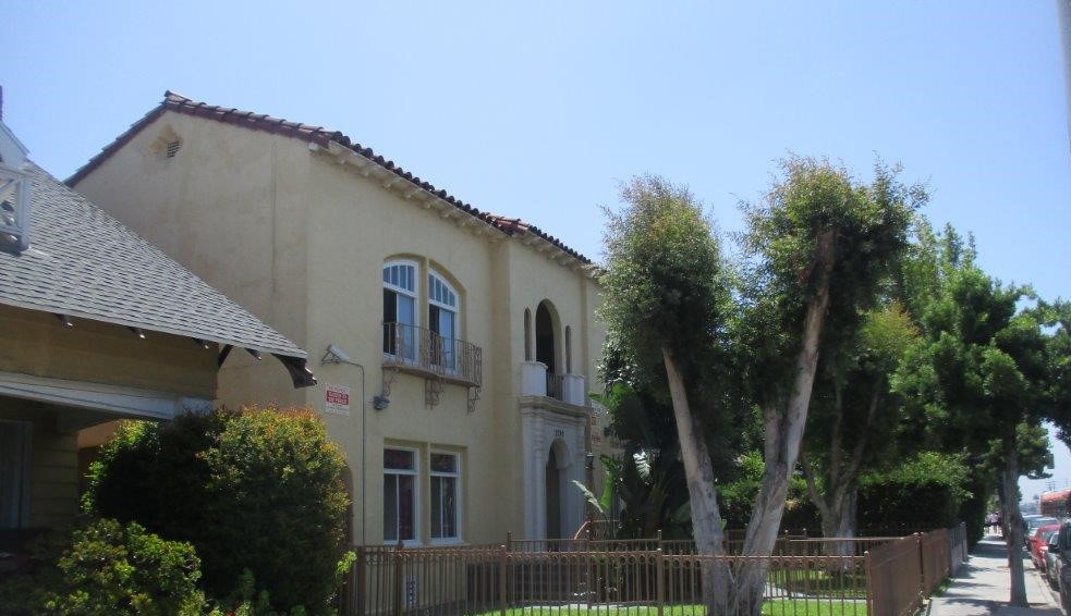 Left side view of a yellow two story building, two windows on first floor on each side, two windows with old fashion iron balcony on each side, wide white pilar in the main entrance door, steps with handrails, gated, multiple trees.