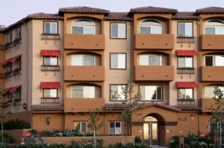 Front side of a two brown tones four story building, multiple windows some with balconies and others with red shades on top, plants and trees all around the building.