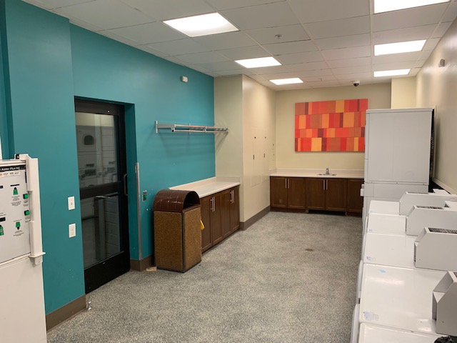 View of a big laundry room, multiple white washers and dryers, brown wood shelves for folding clothes, a clothing rack hanging on the wall, brown wood cabinet with a sink, Big brown trash can, colorful picture frame.
