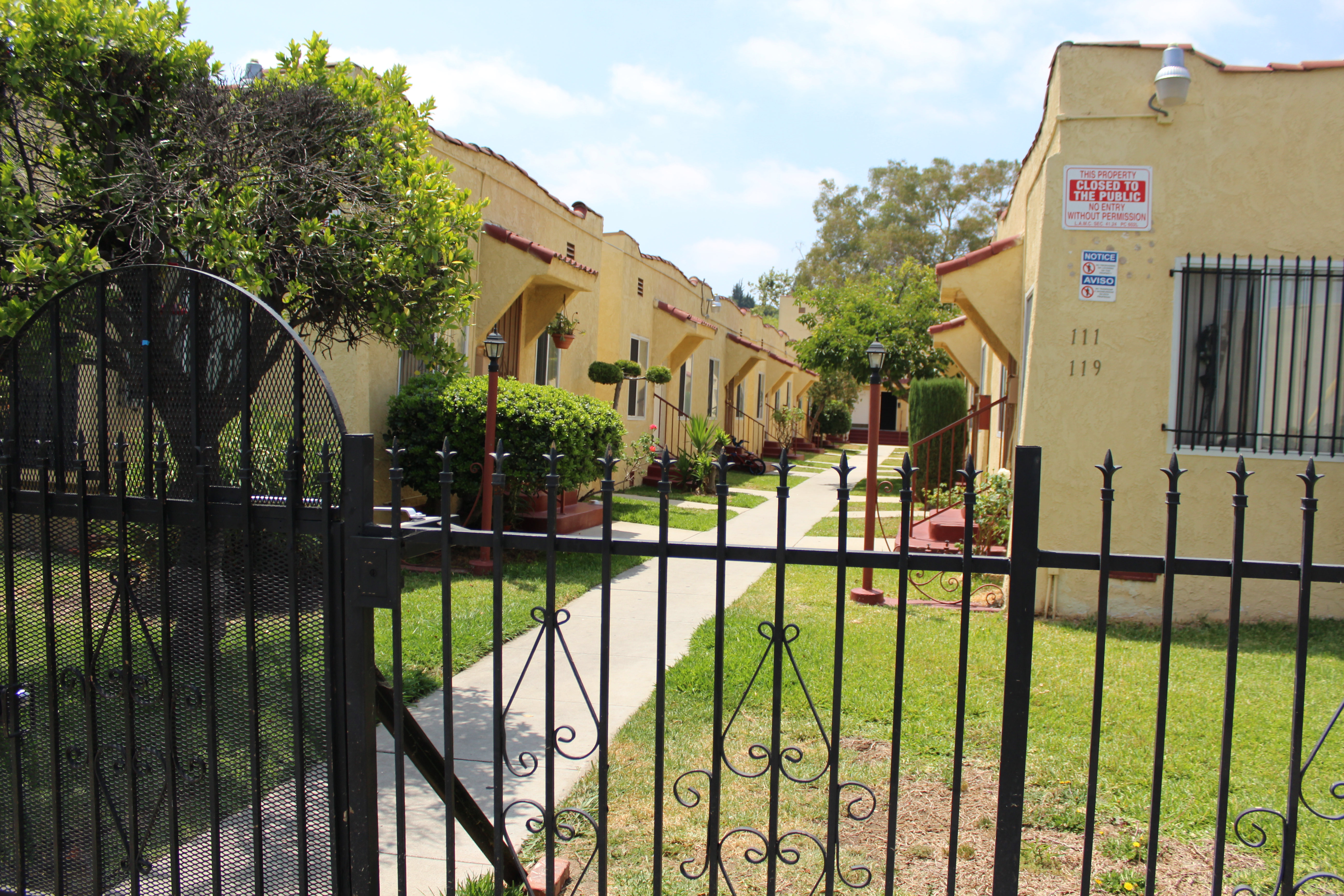 Street view of front entrance to the courtyard style units with a wrought iron gate.  Each unit has a three step exit with handrails, grass sections by each unit.