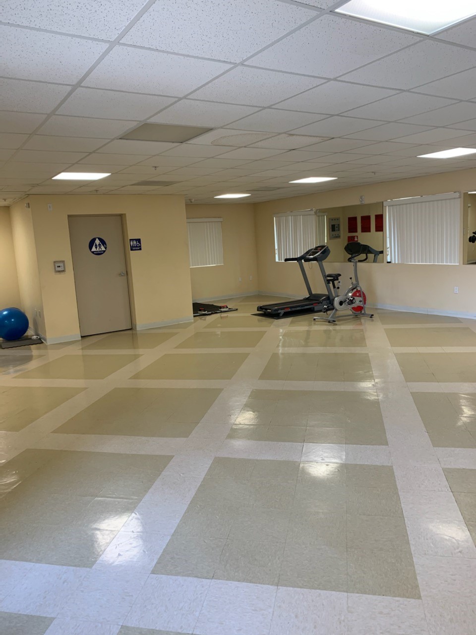 View of a large room with a trreadmill, exercise bike, and a yoga ball. Thre is a gender neutral restroom and the floor is tiled.