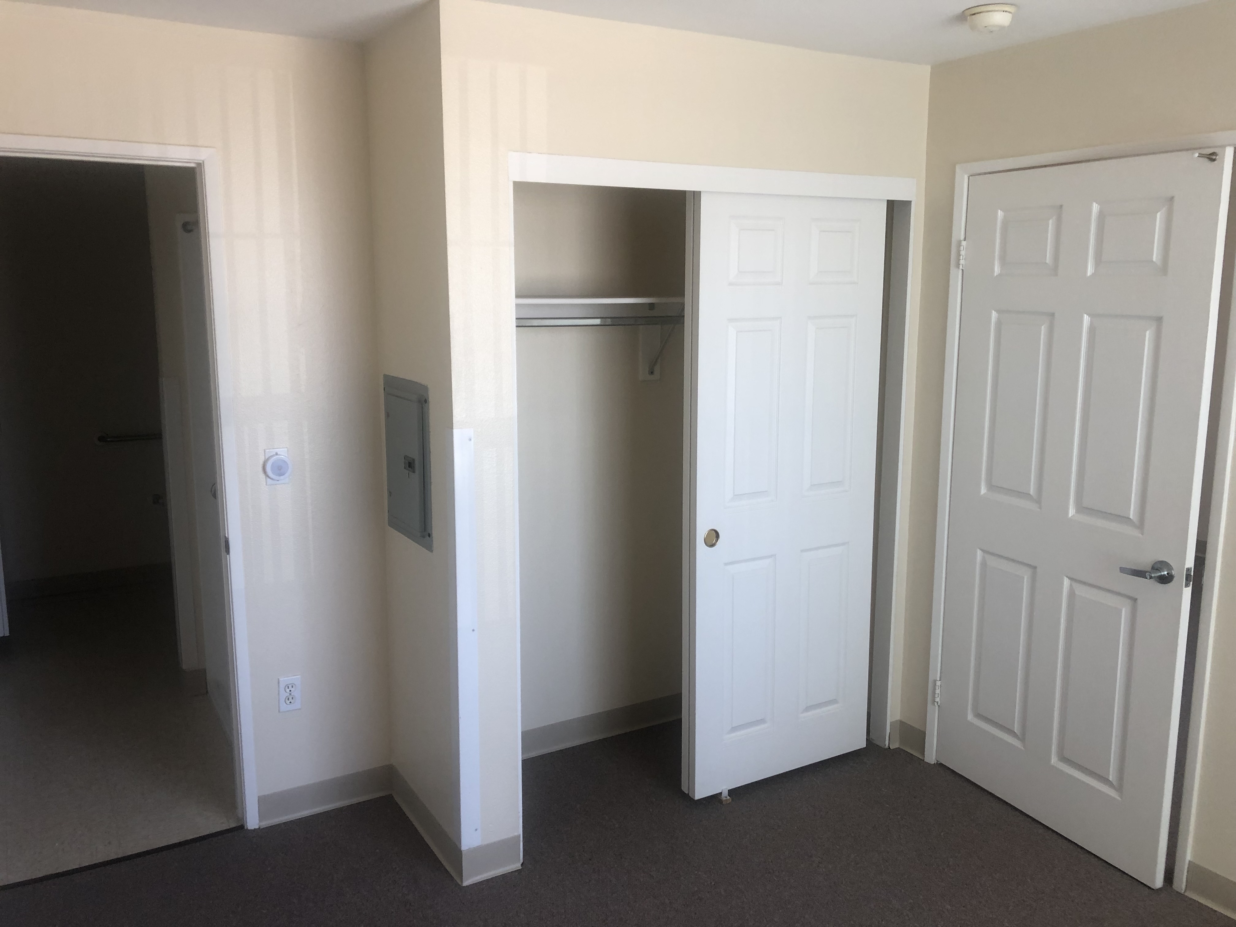 Interior view of unit. Image consists of a small closet with sliding doors, electricity control oanel on wall, and an open door leading to a different room that has a grab bar on the wall.