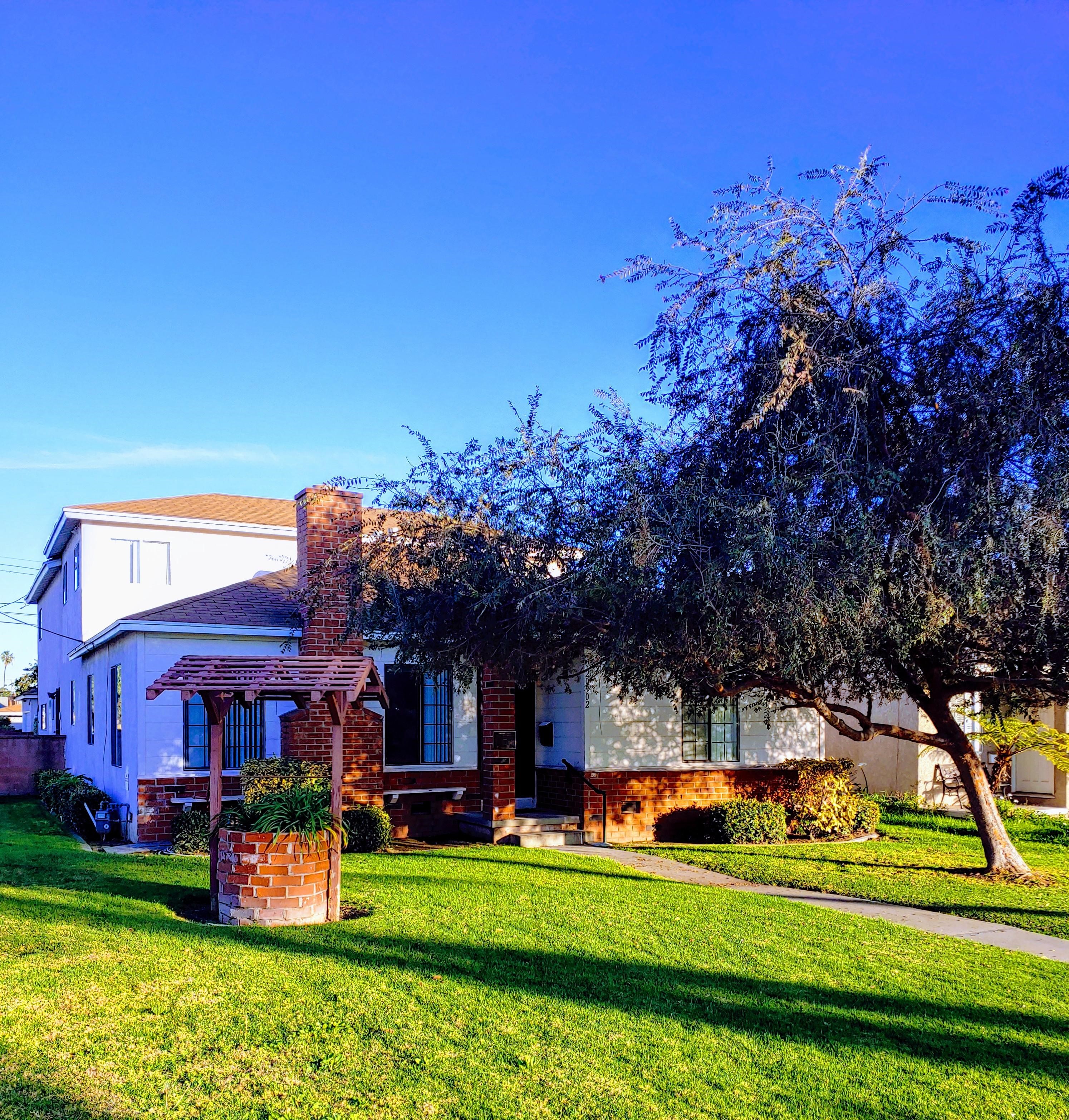 Exterior view of Denker House with a brick chimney, long walkway to entry bordered by a brick well and larg tree on either side.