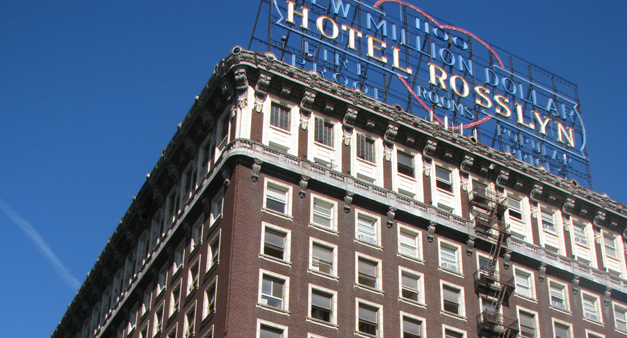 Top view of a large brown building. On the rooftop sits a large sign that reads "Hotel Rosslyn Rooms". Building is old fashioned as is equipped with outter fire escapes.