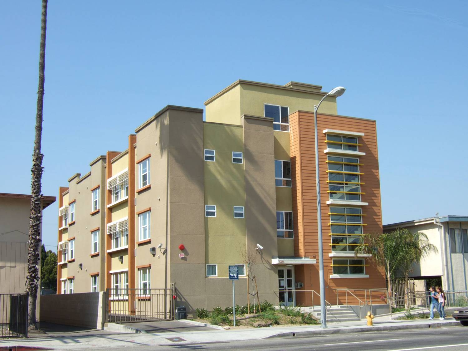 Three story multicolor building. Entry way has stair and ramp access. There is gated parking on premises. Front of the building has a small bush area.